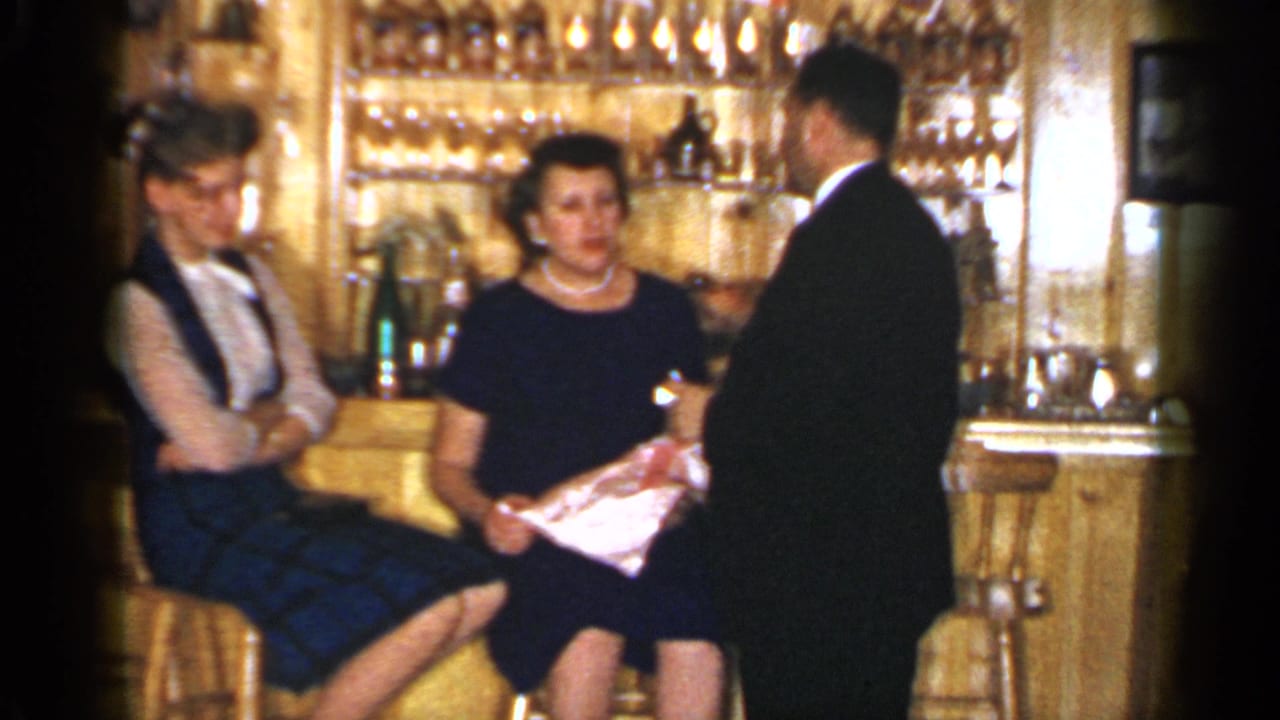 A suited man converses with two seated women at a warmly lit Long Island bar in the 1965 8mm home-movie scene.