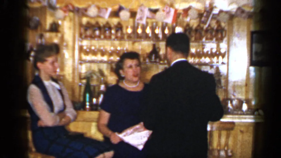 The home-movie frame shows two women at a Long Island bar and a man seen from behind during a 1965 indoor conversation.