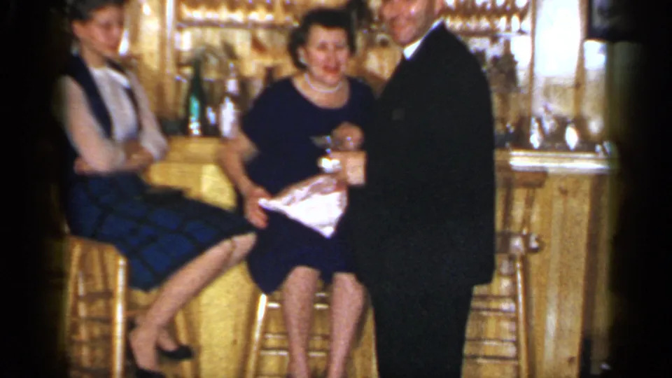 A 1965 Long Island bar scene shows a seated woman smiling while a suited man stands close and another woman sits nearby.