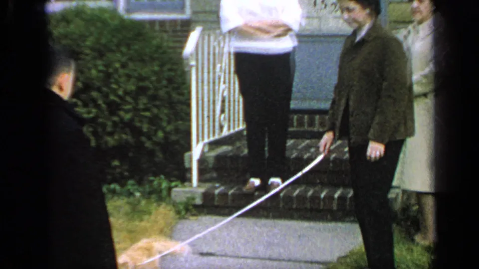1965 Long Island home-movie frame showing adults by a house porch while a ginger cat on a leash walks across the foreground.