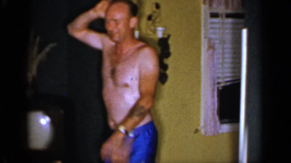 A shirtless adult man in blue swim trunks stands in a private interior, raising one hand near his head beside shutters and a doorway.