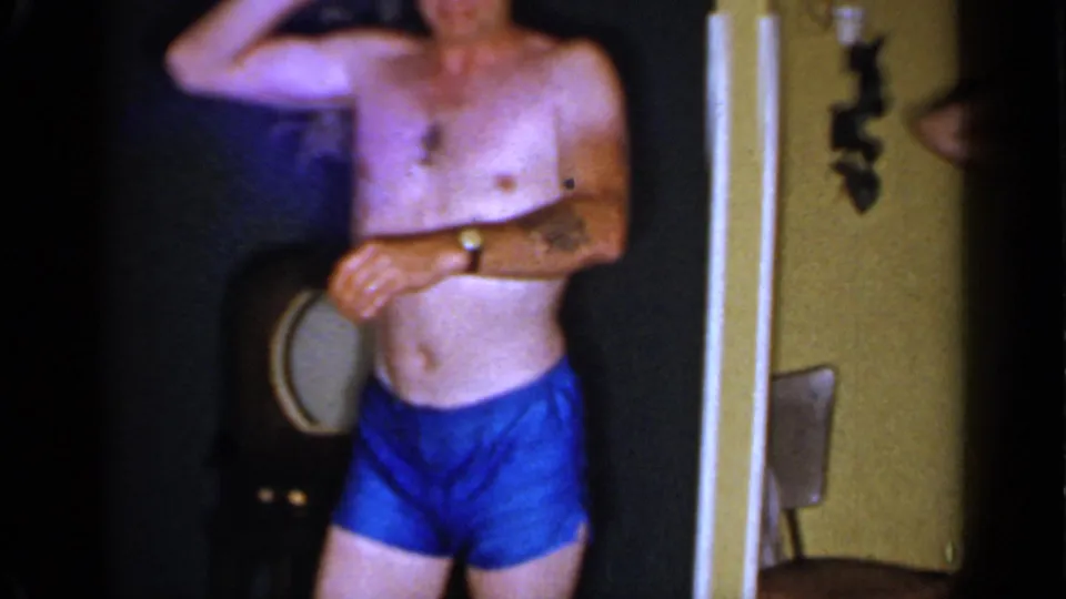 A shirtless adult man in blue trunks stands in a doorway, partially cropped, with household furnishings visible behind him.