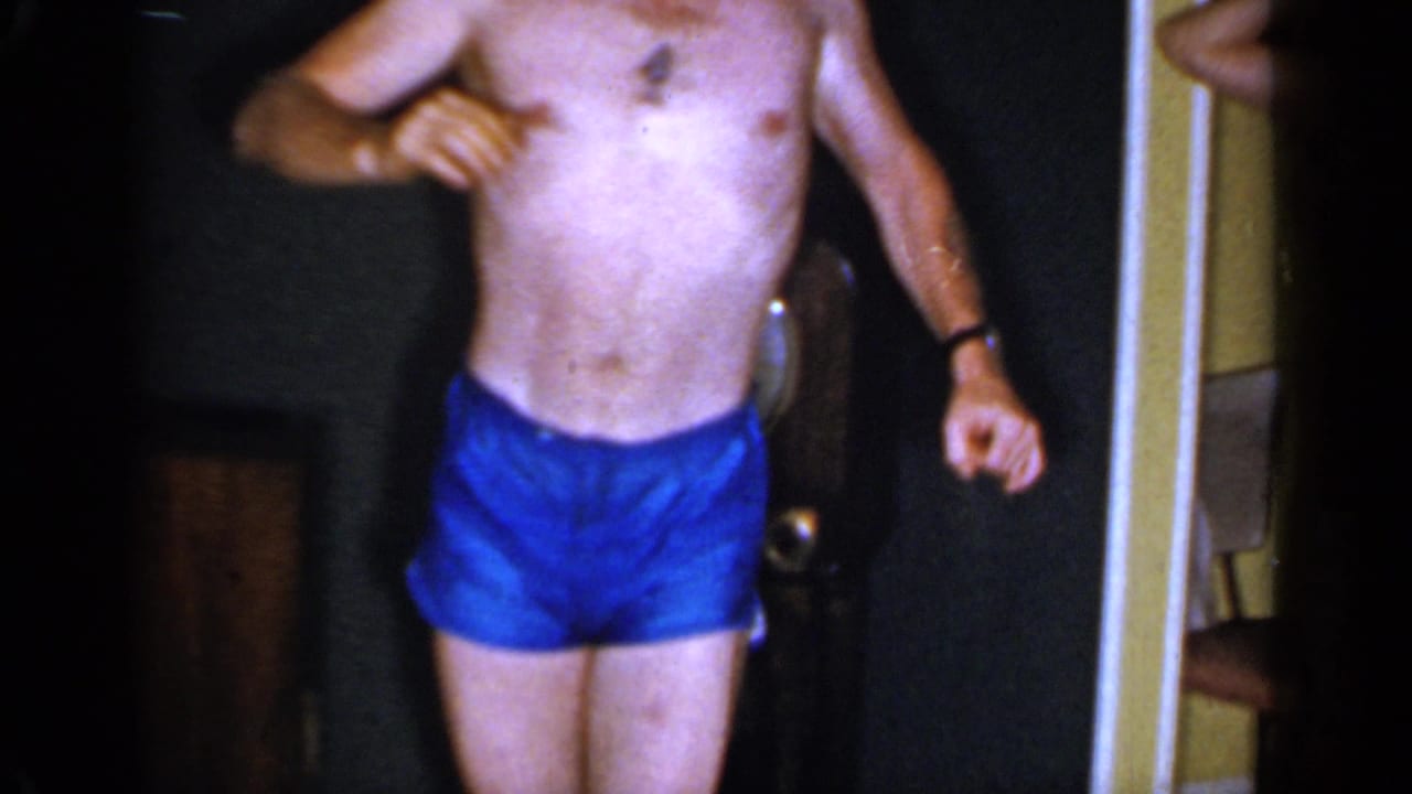 Shirtless man in blue trunks dancing barefoot inside a dim room.