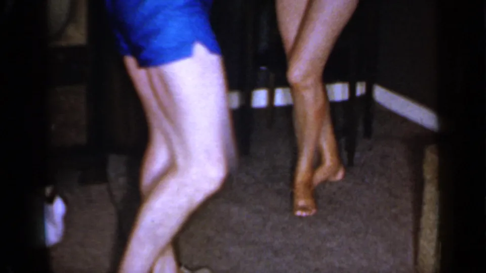 A low-framed 1966 home-movie view shows the barefoot man dancing beside the woman, with carpet and dark furniture visible behind them.