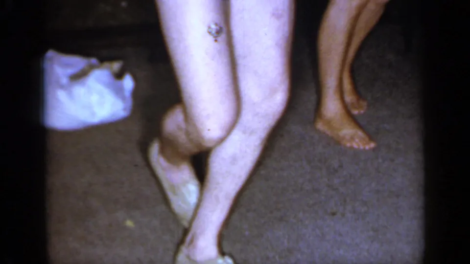 The frame catches the man's crossed barefoot step near the woman in a dark dress, with a white bag on the carpet in the private interior.