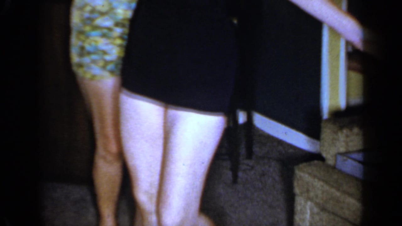 Two women in short dresses are framed from the waist down in a Mineola interior, with sofa edge and steps nearby.