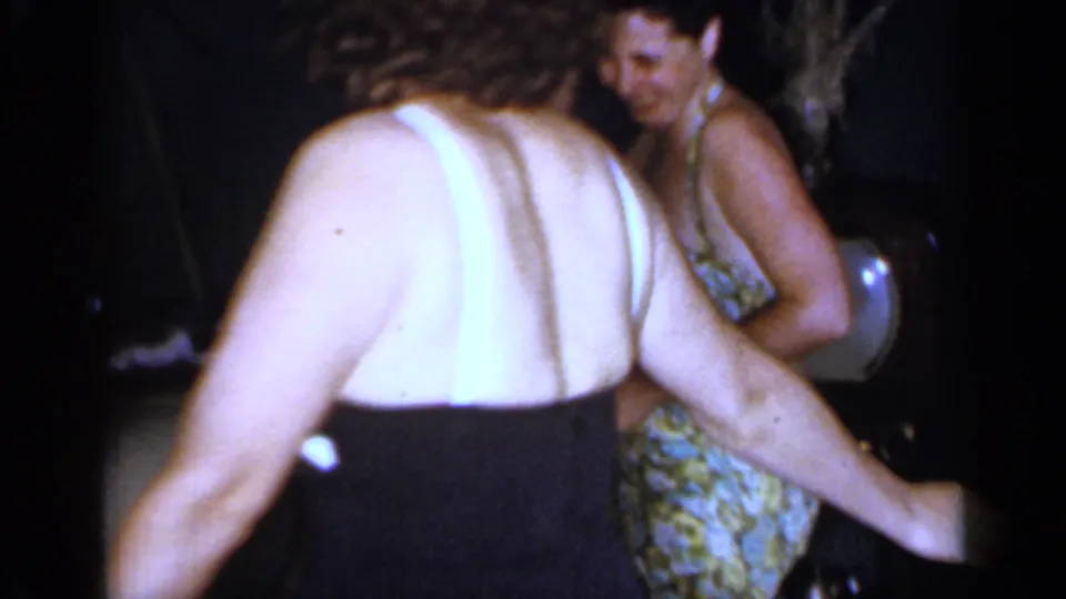 1966 Mineola home-movie frame showing two women in swimsuits near a dark background during a casual summer gathering.