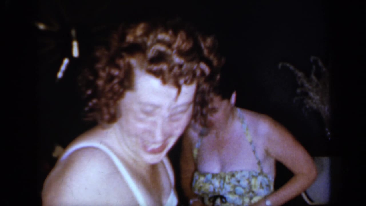 A woman in a light-strap swimsuit laughs beside another woman in a floral swimsuit.
