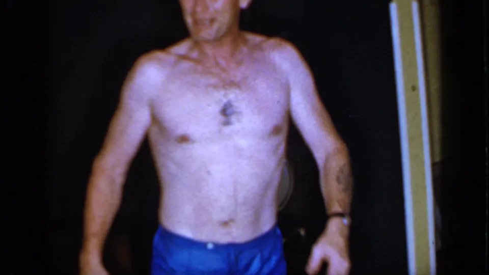 1966 Mineola frame showing a shirtless man in blue swim trunks standing near a dark entry or wall.