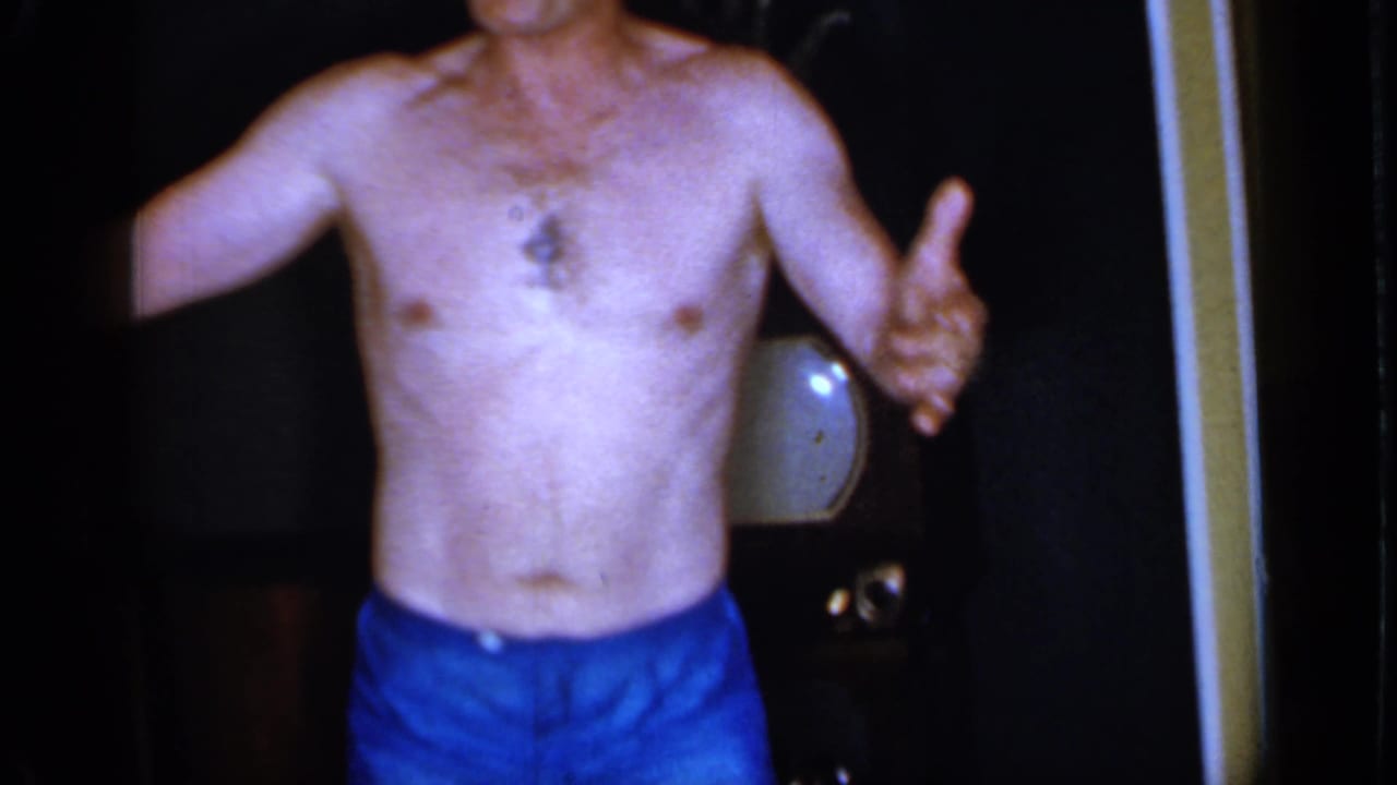 Shirtless man in blue shorts stands in a dim room near a television set.