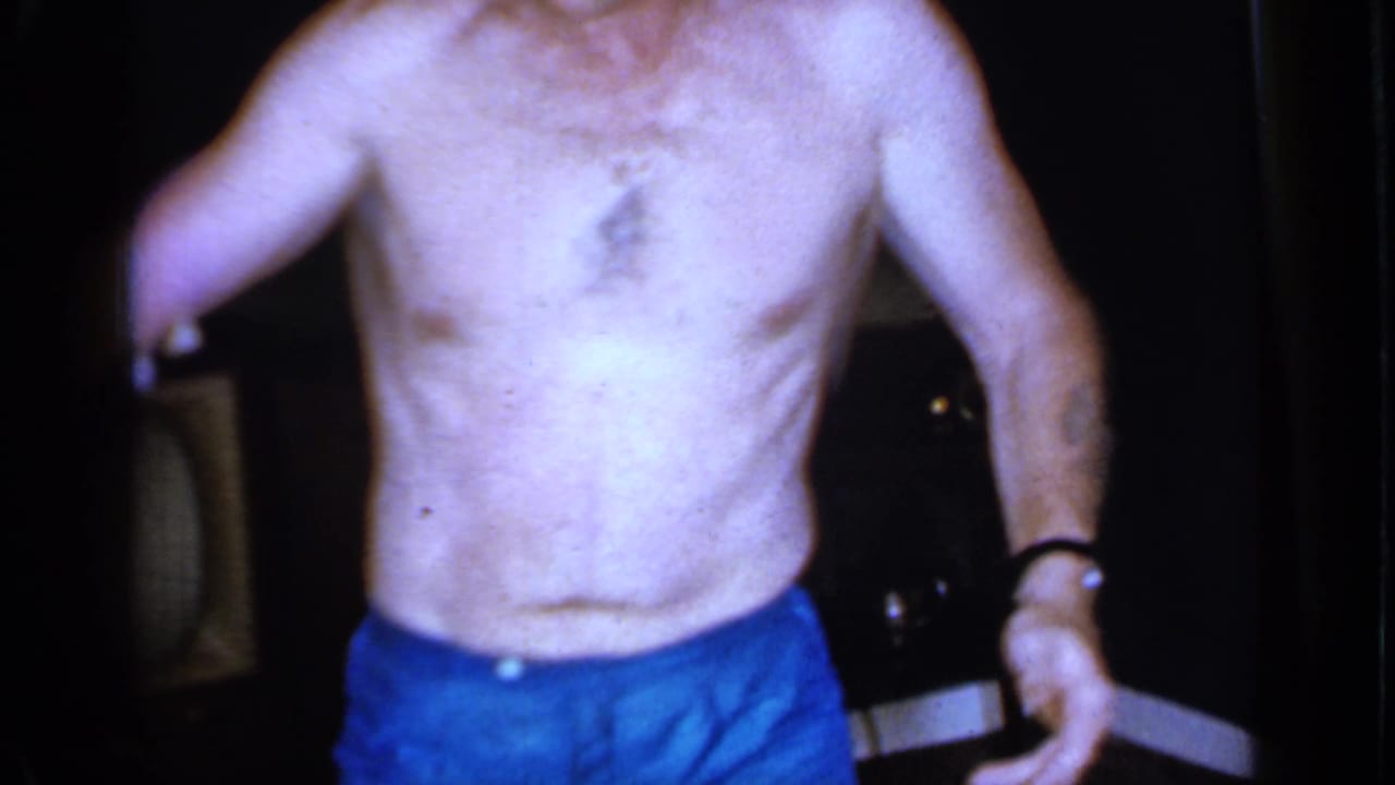 Shirtless man in blue shorts stands closer to a dark television and furniture.