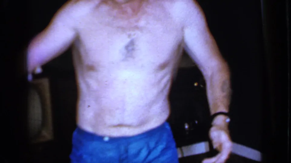 The man remains framed from neck to shorts in the Mineola interior, with a dark television set and furniture visible behind him.
