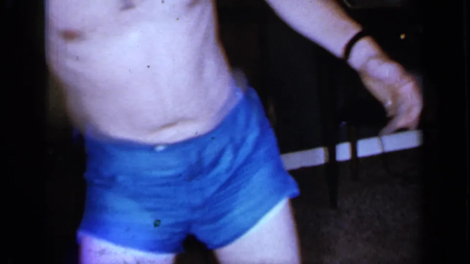 A closer cropped frame shows the man’s torso, blue shorts, wristwatch, and part of the dim private room around him.