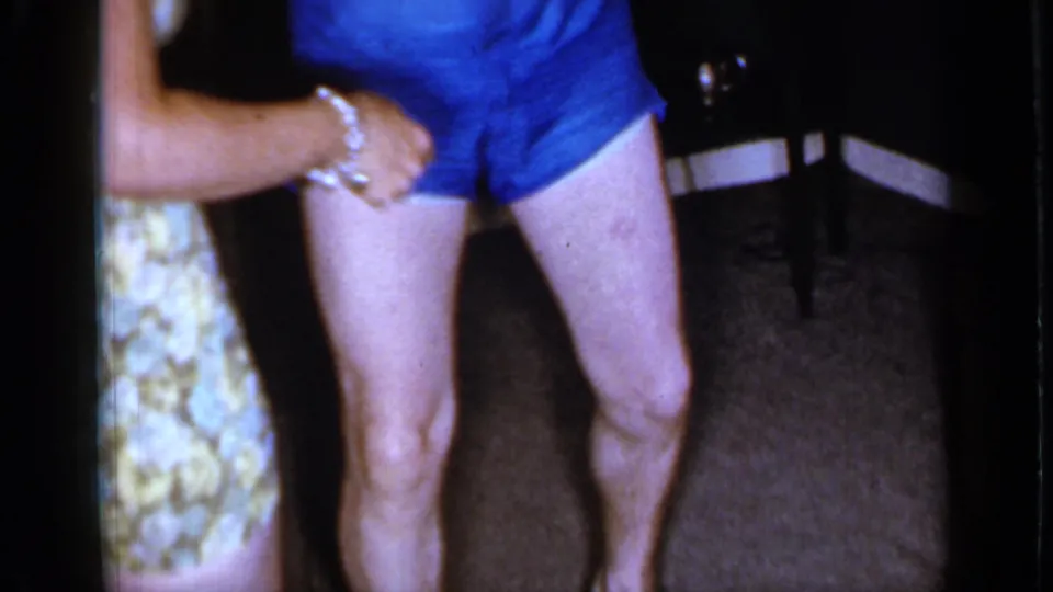 A woman in a floral dress briefly enters the 1966 Mineola frame, reaching toward the shirtless man standing on the carpet.