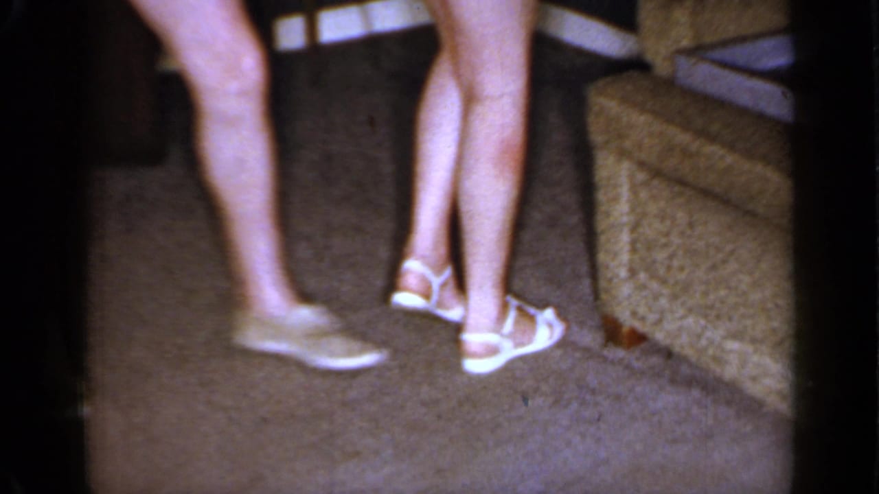 Two adults' legs and sandals near a textured sofa on carpet.