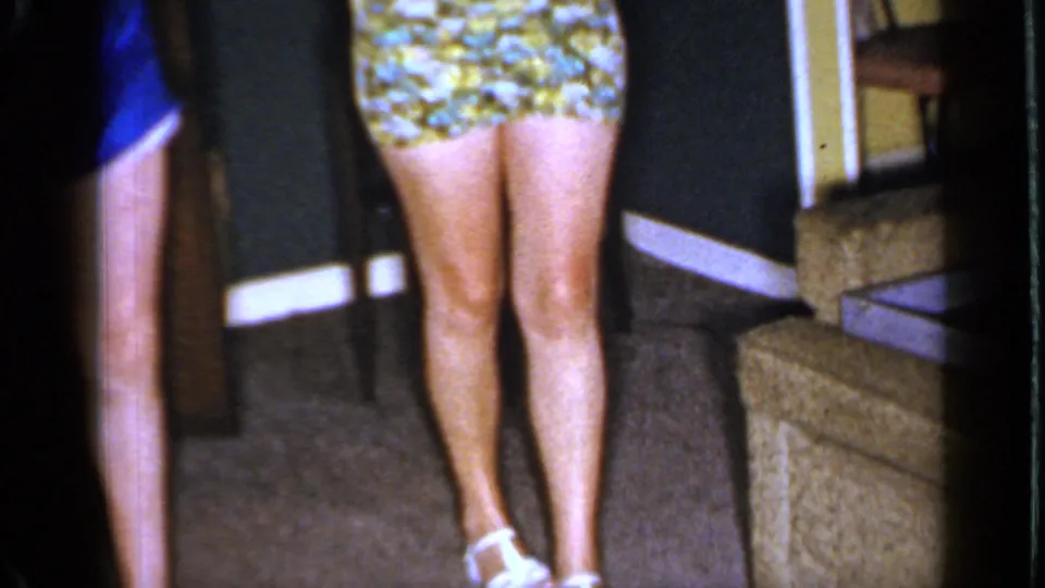 1966 Mineola living room frame showing a floral mini dress, adult legs, carpet, dark walls, and upholstered furniture.