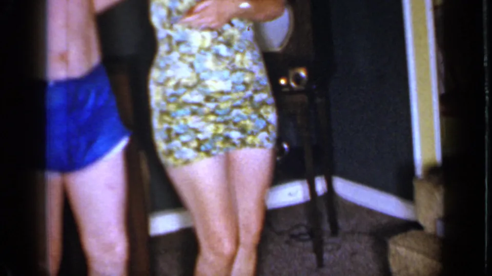 1966 Mineola home-movie frame showing a floral mini dress, blue swim trunks, carpet, dark walls, and a sofa in a private interior.