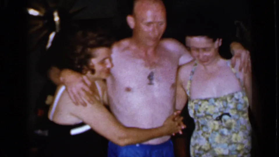 A 1966 Mineola home-movie frame shows a shirtless man standing between two women in bathing suits, with their arms close around one another.