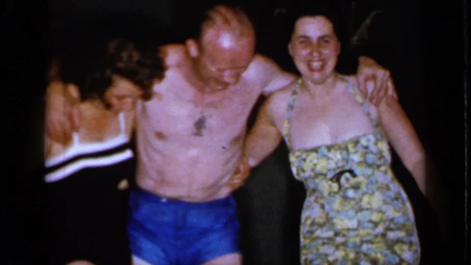 In 1966 Mineola home-movie footage, a shirtless man in blue trunks stands between two women in swimsuits as they smile together.