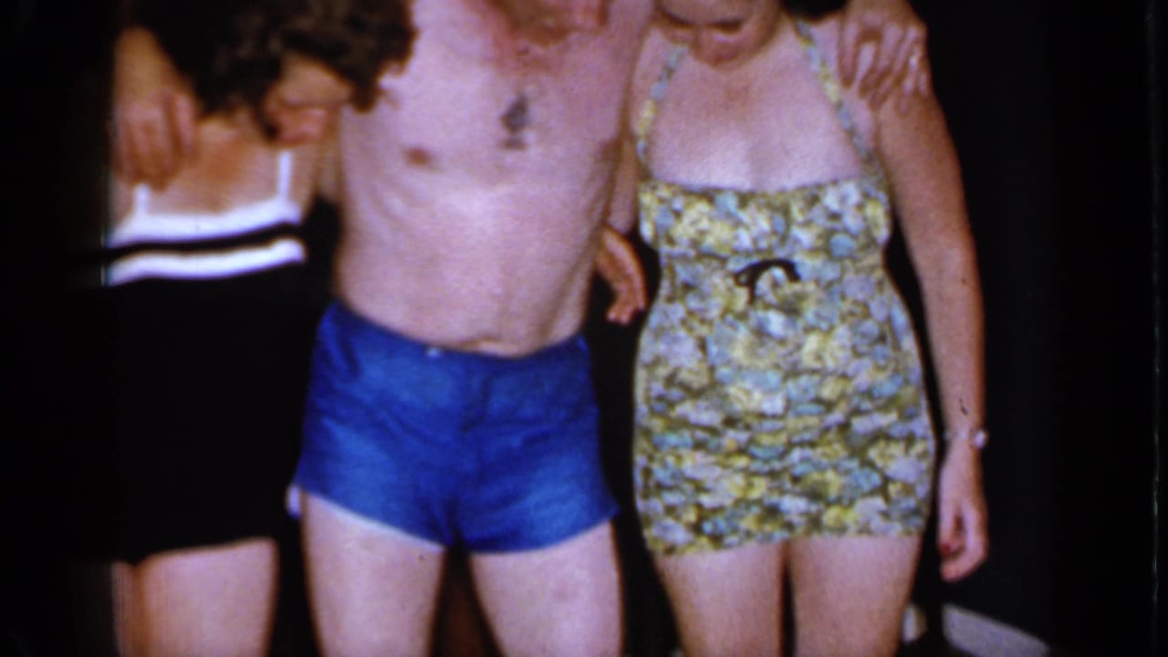 A close 8mm frame shows the linked adults in swimsuits, with the central man in blue trunks and a floral swimsuit at right.