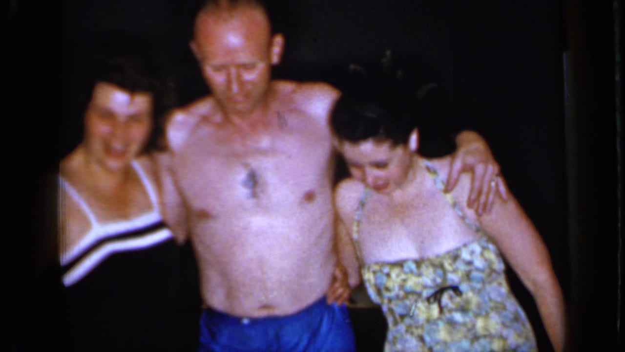 1966 Mineola frame with a shirtless man in blue trunks standing between two women in swimsuits during a casual indoor gathering.
