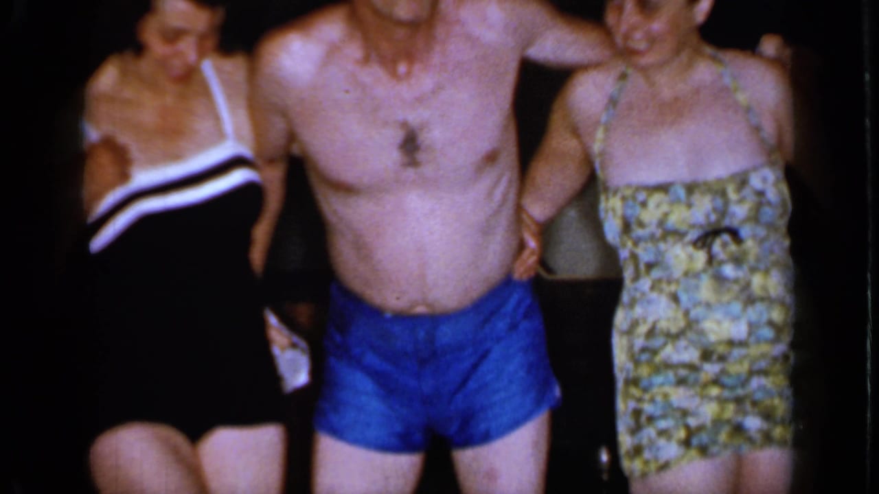 Shirtless man in blue swim trunks stands between two women in swimwear.