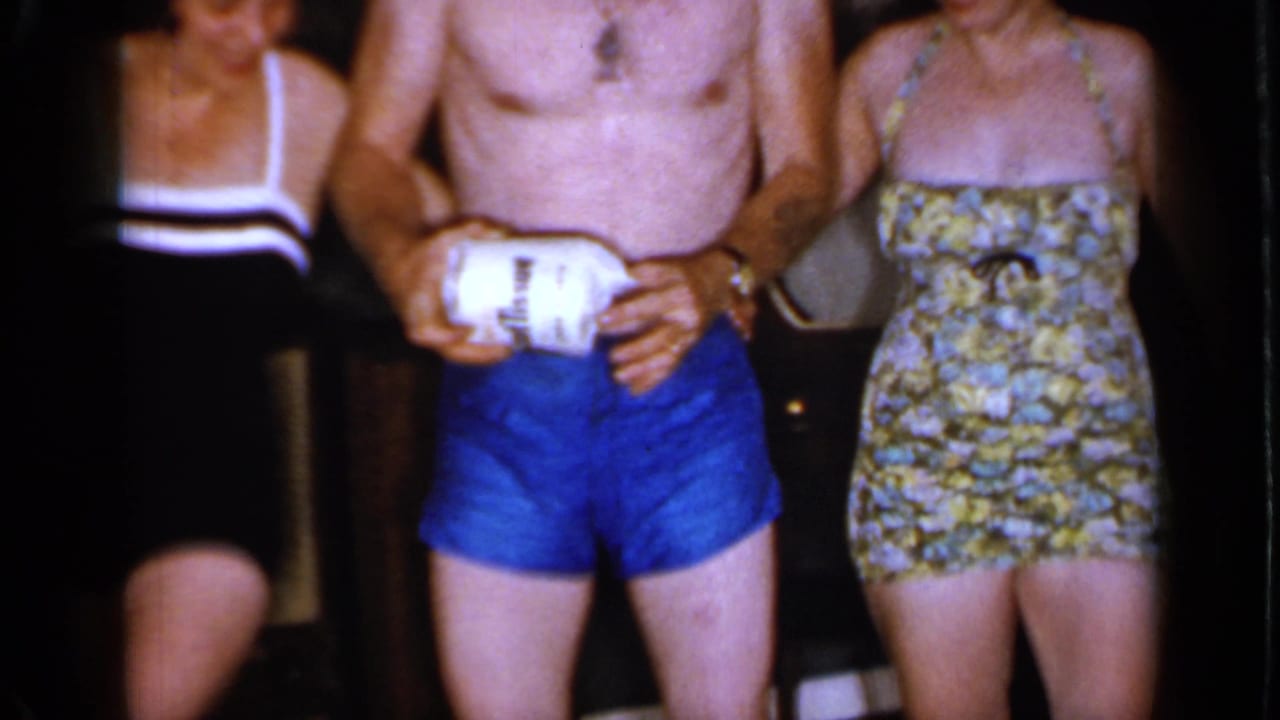 Man in blue swim trunks holds a toilet paper roll beside two women.