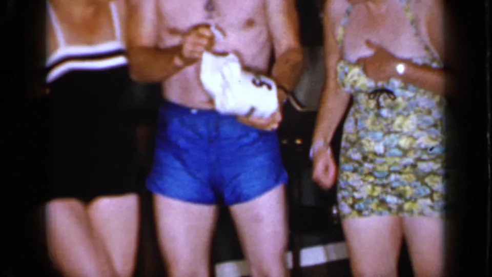 A close handheld frame shows the man handling a toilet paper roll while the women stand close by in vintage swimwear.