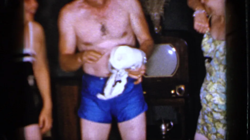 The man holds the loosened toilet paper roll in front of him, with a woman partly visible and chairs in the dim private setting.