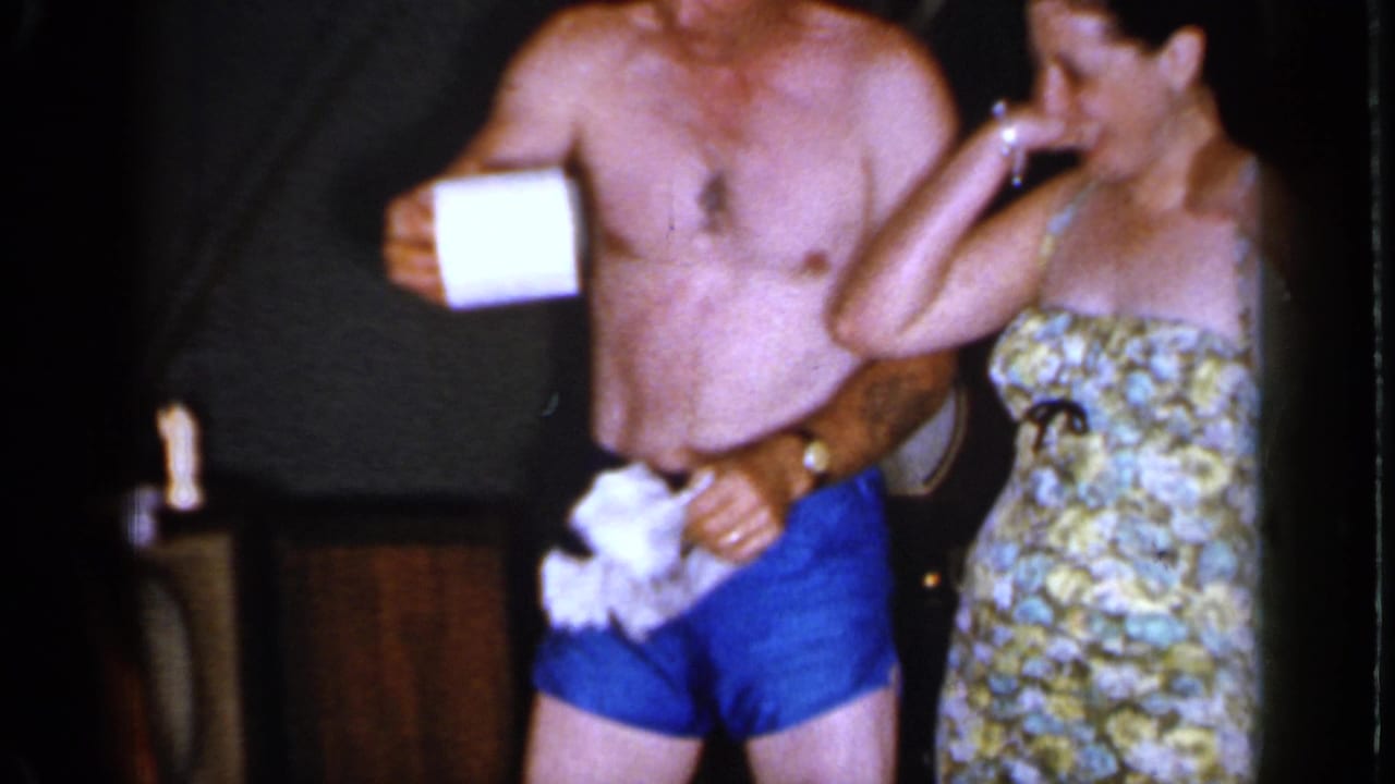 Adults in bathing suits stand indoors near a television while a shirtless man holds toilet paper.