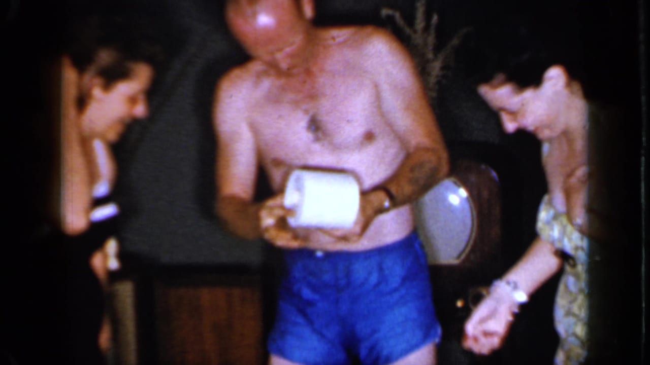 Adults in bathing suits stand in a dim Mineola interior as the man in blue trunks examines or handles a roll of toilet paper.
