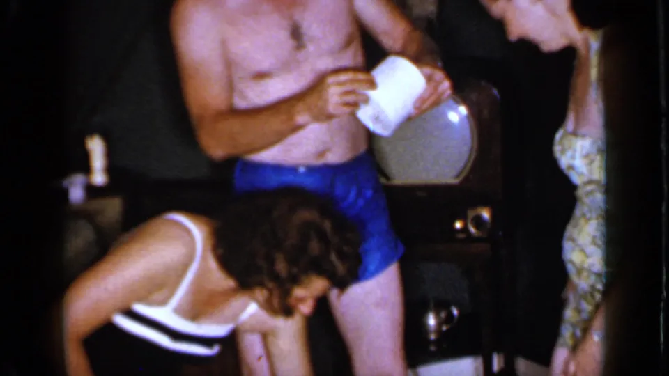 In the 1966 home-movie scene, a woman bends at frame edge while a shirtless man and another woman stand near the television.