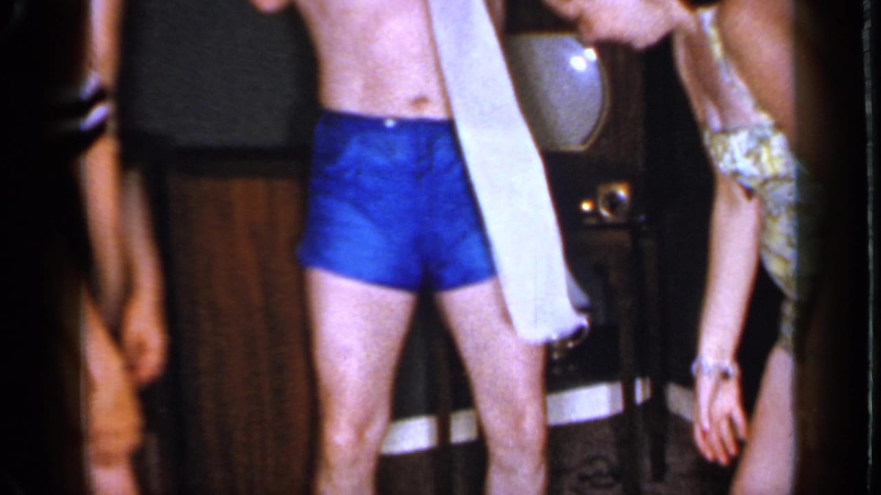 Shirtless man in blue swim trunks holds toilet paper beside women at an indoor party.