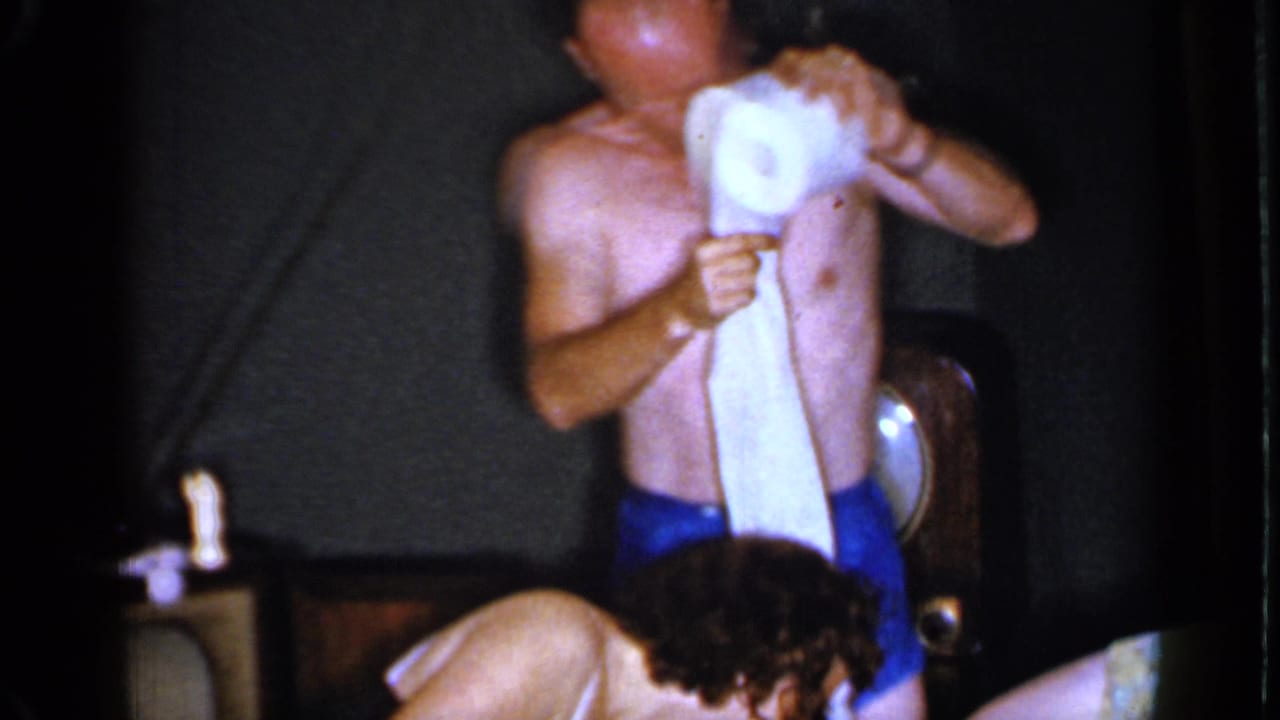 The man in blue trunks holds a toilet paper roll and strip as another guest bends forward in a dim 1960s interior.