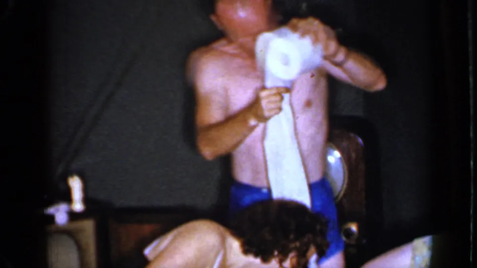 The man in blue trunks holds a toilet paper roll and strip as another guest bends forward in a dim 1960s interior.
