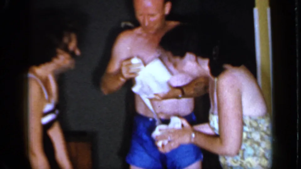 Adults at the Mineola party laugh and gather around the shirtless man as he handles toilet paper in the dim room.