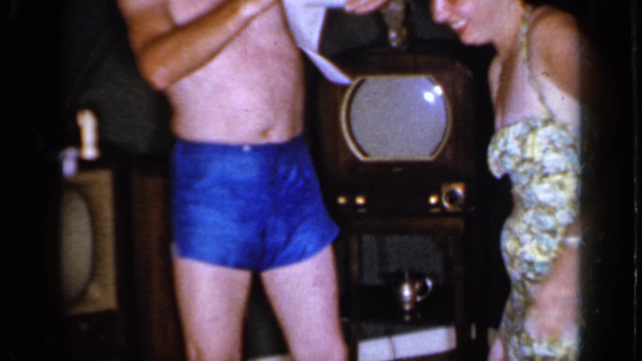 Man in blue swim trunks stands beside woman in patterned swimsuit near a television.