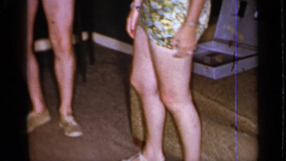 Mineola 1966 home-movie frame showing adults in swimwear from the waist down beside furniture and a household floor.