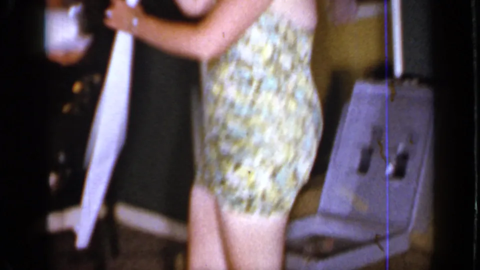 1966 Mineola interior frame with a woman in a patterned swimsuit near a chair, tissue paper, and dark furniture.