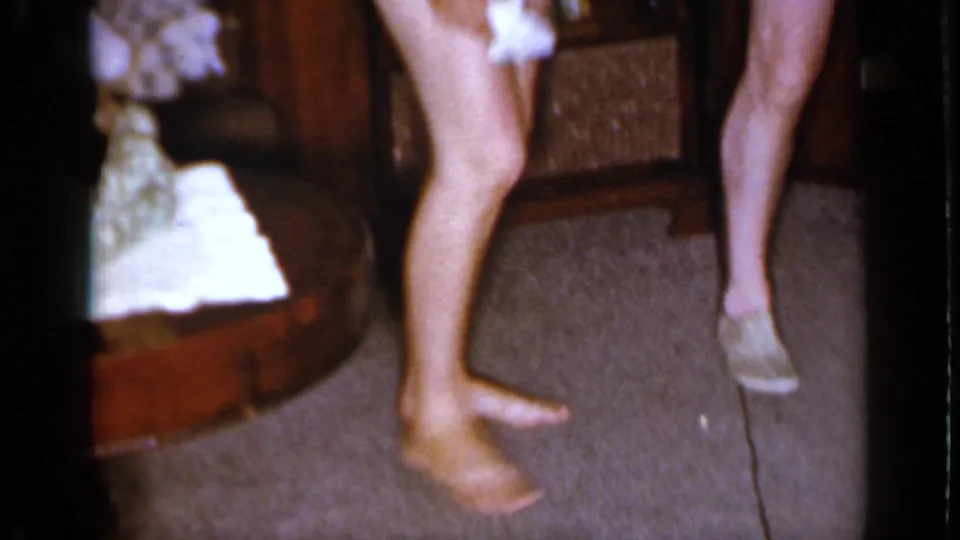 A low, handheld 1966 home-movie frame shows adults' legs and bare feet on carpet as white tissue paper is handled nearby.