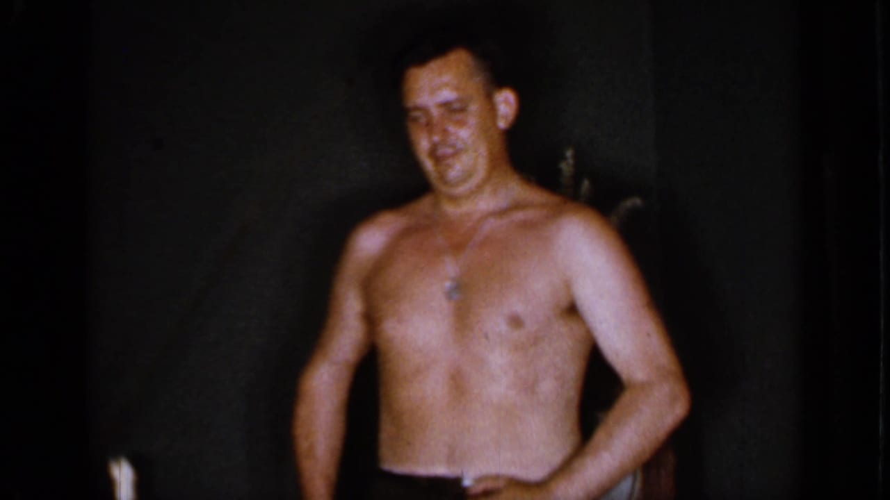 Shirtless adult man in dark trousers standing in a dim interior.