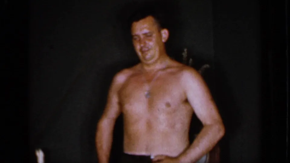 In 1966 Mineola home-movie footage, a shirtless adult man with a necklace faces the camera in a dark room.