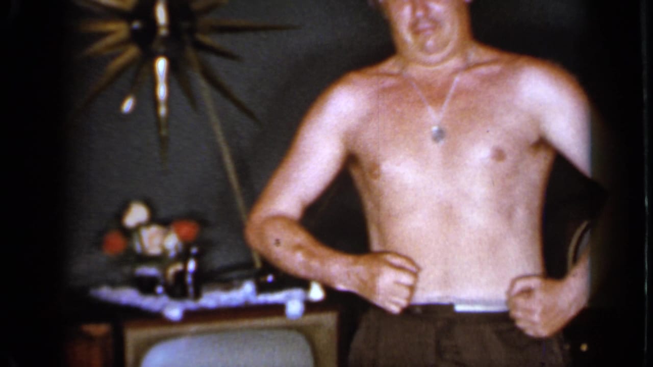 Shirtless adult man holding his waistband near a table and wall decoration.