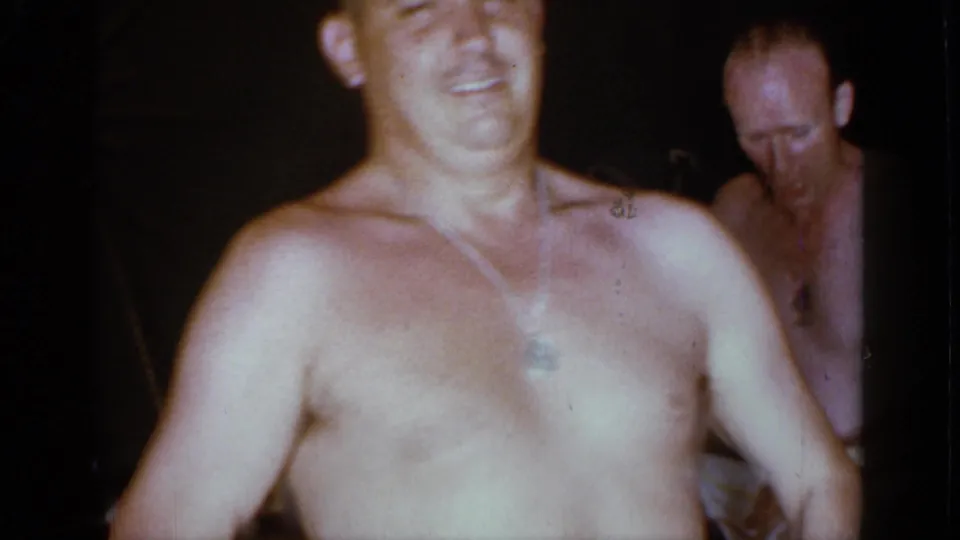 A close shirtless man fills the frame while a second shirtless adult man appears behind him in the dim Mineola room.