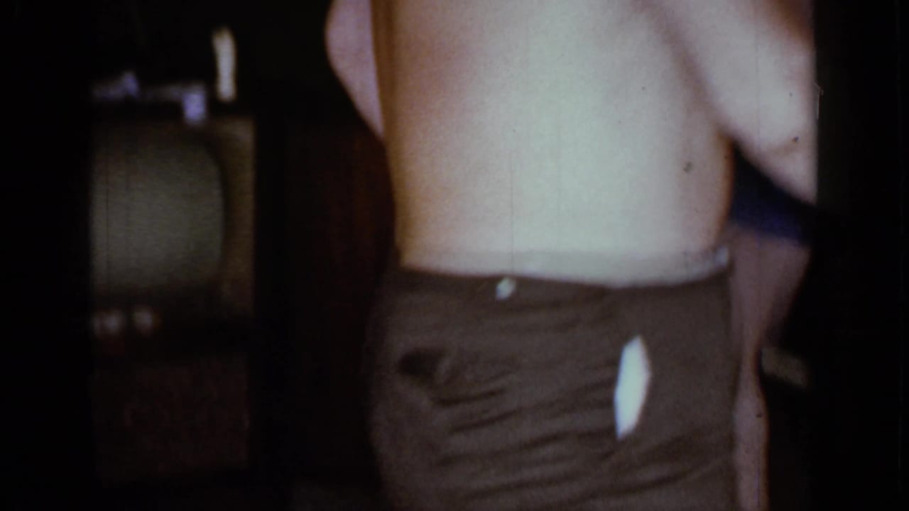 Shirtless man partly framed beside a vintage television in a dim living room.