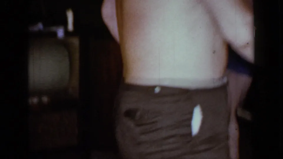 In 1966 Mineola home-movie footage, a shirtless adult man is partly framed near a vintage television inside a dim living room.