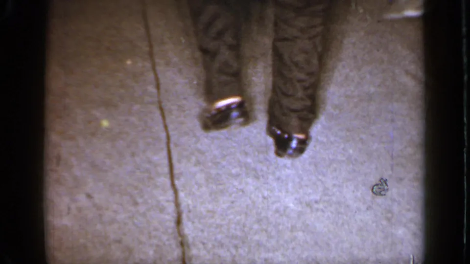 The camera points down toward carpeted flooring, showing a person's lower legs and a small object on the living room floor.
