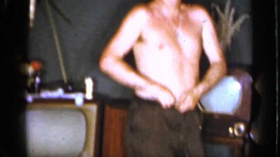A shirtless adult man stands in a Mineola living room, adjusting his pants beside a vintage television and wood furniture.