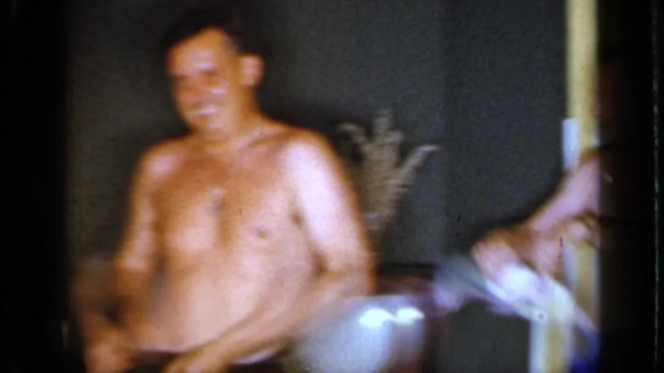 A smiling shirtless adult man dances in the 1966 Mineola living room while holding a white cloth, with another person partly visible nearby.
