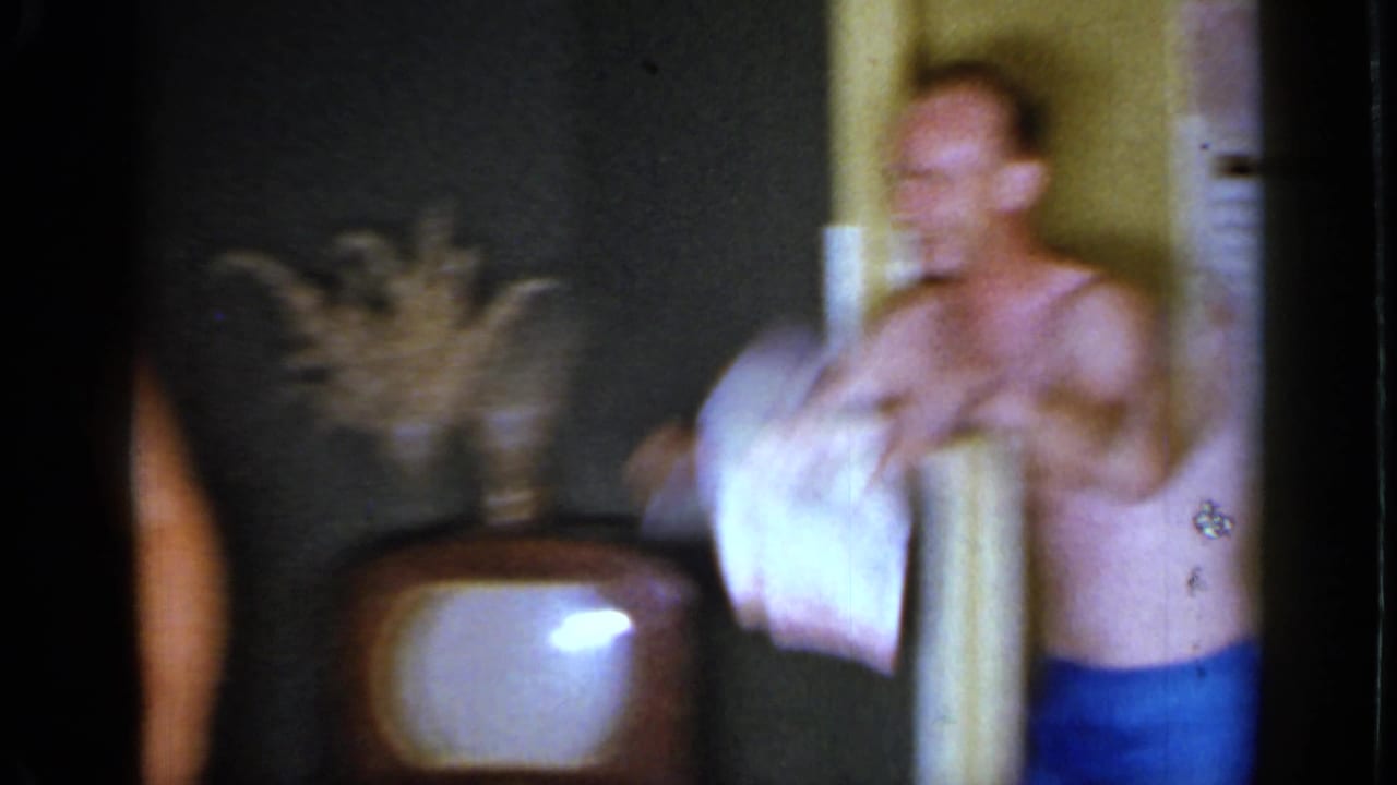 Shirtless man moving in a dim living room beside a wood cabinet television.